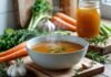 Bone Broth Benefits for Health, Skin, and More A steaming bowl of bone broth on a wooden table surrounded by fresh vegetables and herbs in a bright kitchen.